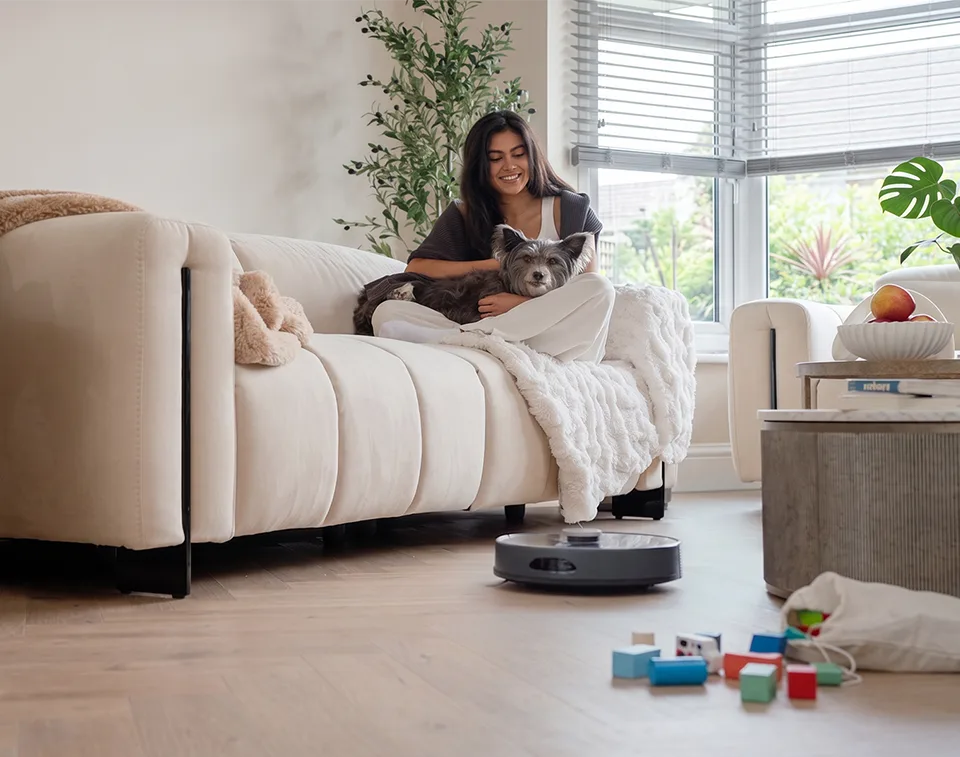 Robot vacuum cleaner Vacmaster REi vacuuming in front of sofa with woman and dog
